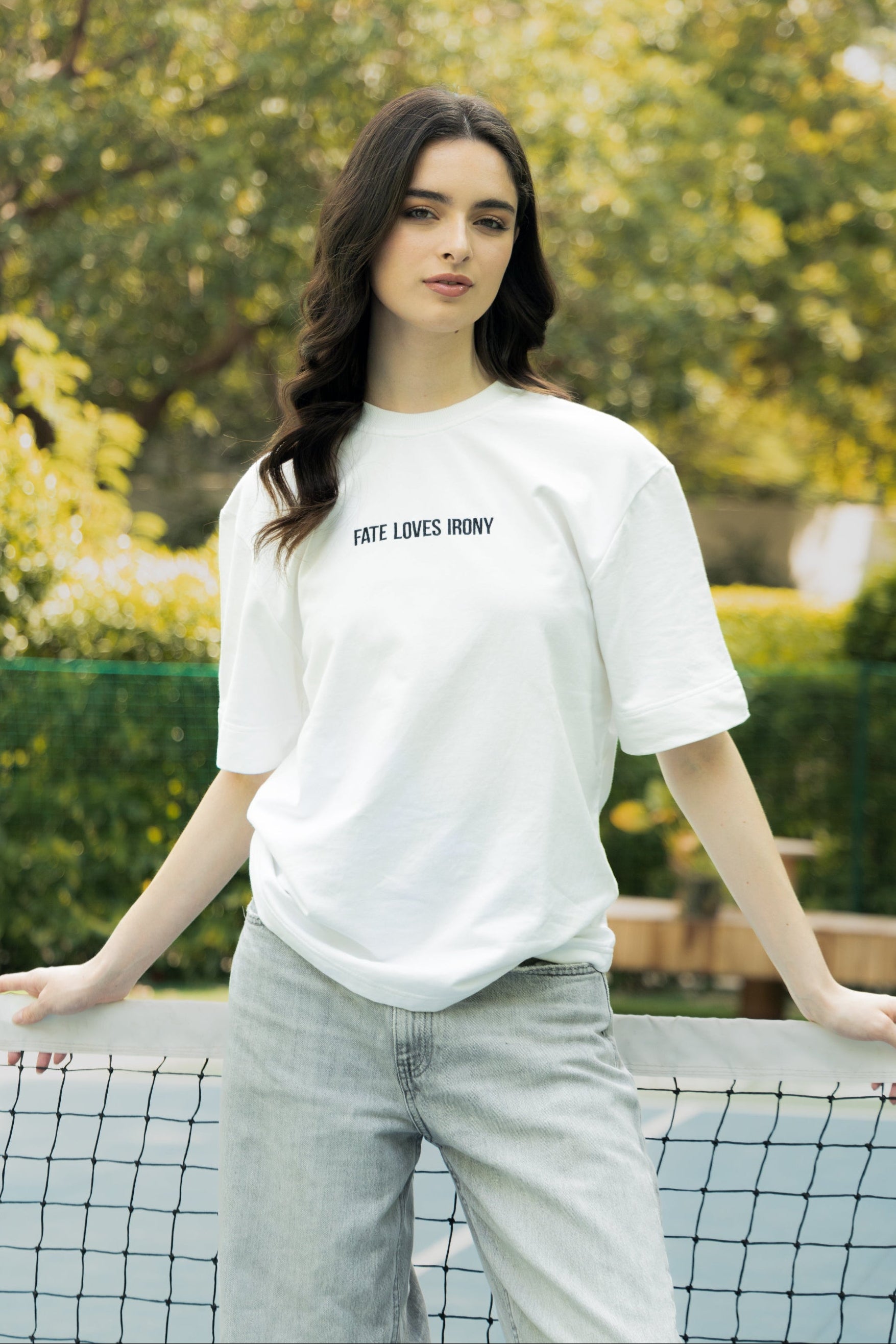 Woman wearing a white t-shirt with text, standing on a tennis court with greenery in the background